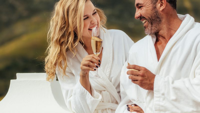 Cheerful couple in bathrobe talking and laughing in the balcony with glass of wine. Man and woman in bathrobes spending time at together.